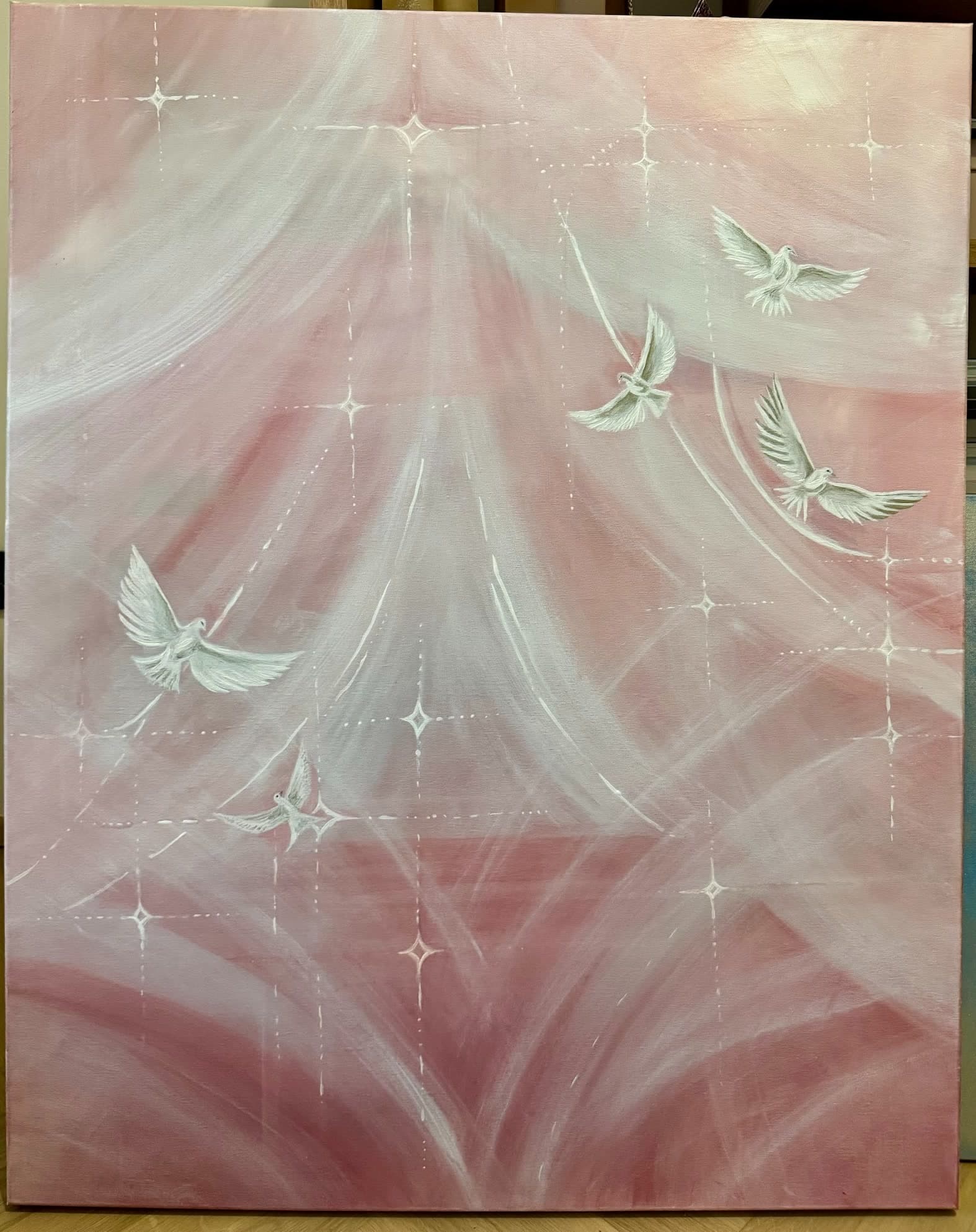 Photograph of an original painting by Urška depicting four white doves in flight across a soft pink and rose-gold sky, with light rays and small sparkling stars.